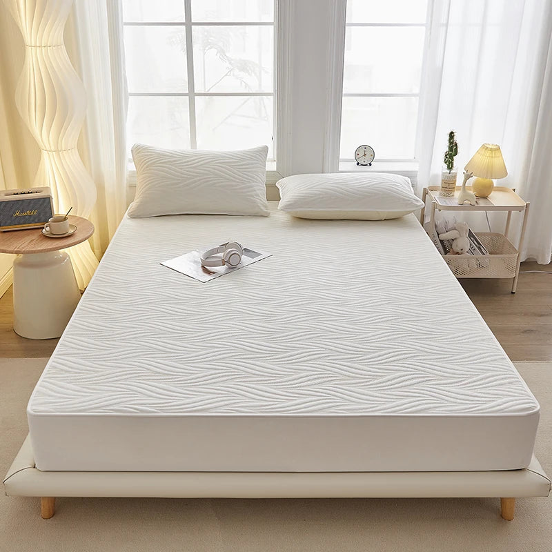 A white fitted mattress protector is displayed on a bed with pillows and natural light.