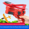 A hand holds a red foam glider aircraft gun toy, ready to launch, with an orange airplane visible nearby.