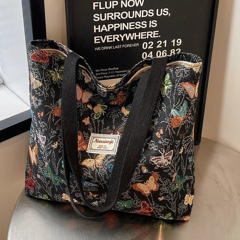 A black corduroy tote bag with floral accents is displayed in front of a sign promoting happiness.