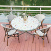 A round table set for outdoor dining featuring a floral tablecloth, white chairs, and greenery by a body of water.