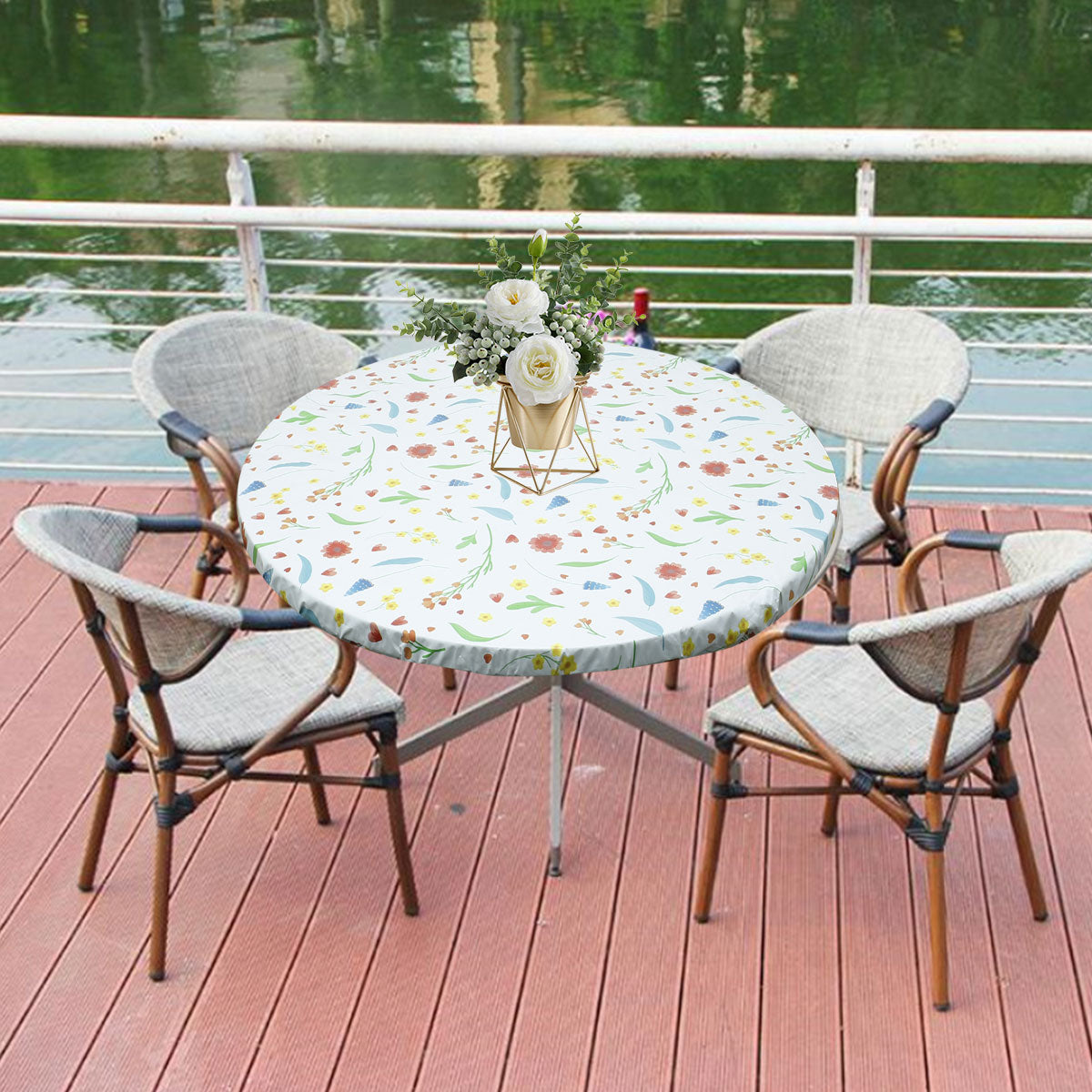 A round table set for outdoor dining featuring a floral tablecloth, white chairs, and greenery by a body of water.