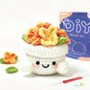 A white crocheted animal figure with a smiling face sits next to a purple bag labeled as a DIY crochet kit.