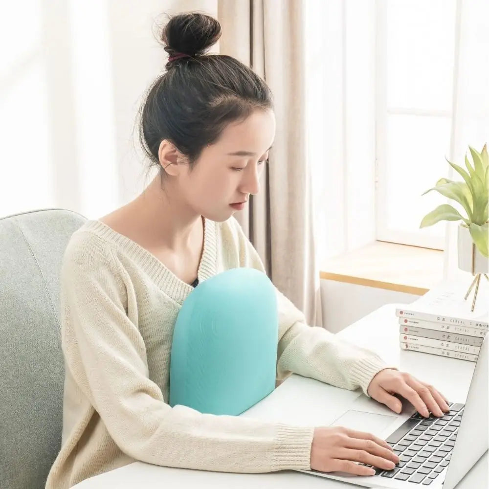 A woman is using a laptop while resting her head on a light blue memory foam travel neck pillow.