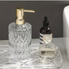 A clear glass soap dispenser with a gold pump and a small dark bottle on a glass tray.