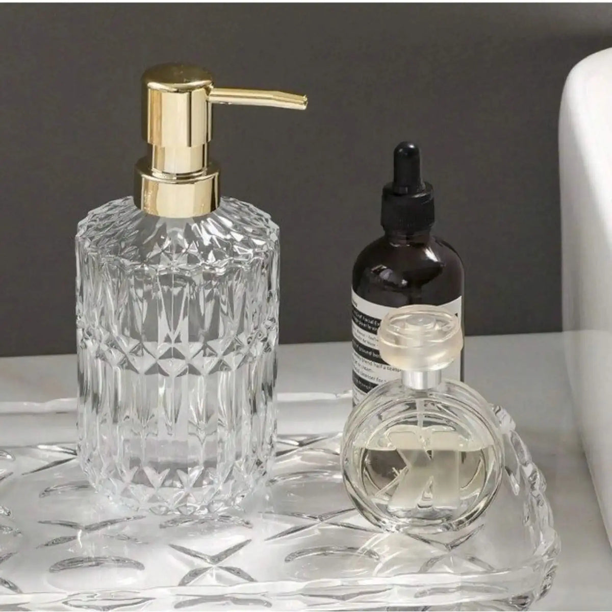 A clear glass soap dispenser with a gold pump and a small dark bottle on a glass tray.