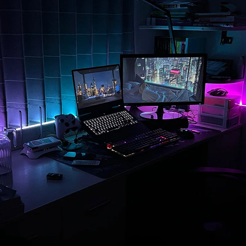 A dark desk setup featuring a laptop, monitor, keyboard, and various electronic components illuminated by purple and blue RGB