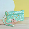 A hand holds a mint green daisy print PU leather wristlet wallet with a gold zipper closure against a yellow background.
