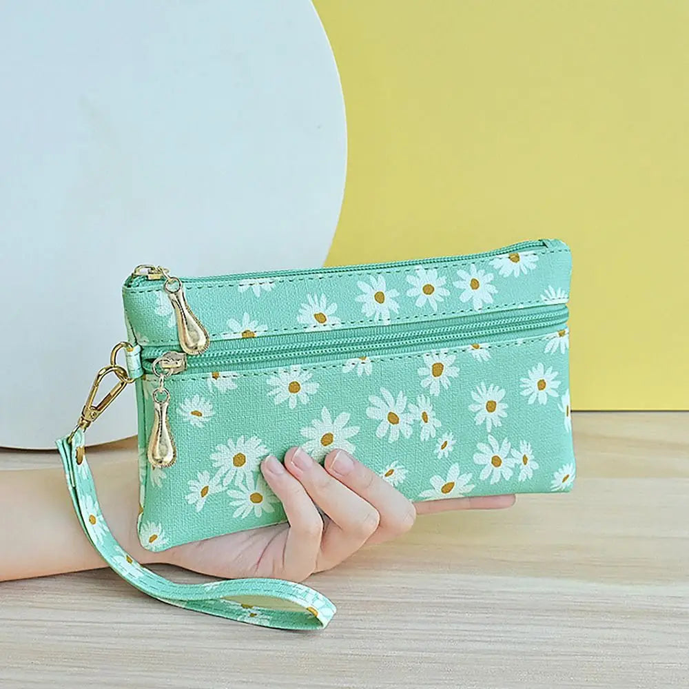 A hand holds a mint green daisy print PU leather wristlet wallet with a gold zipper closure against a yellow background.