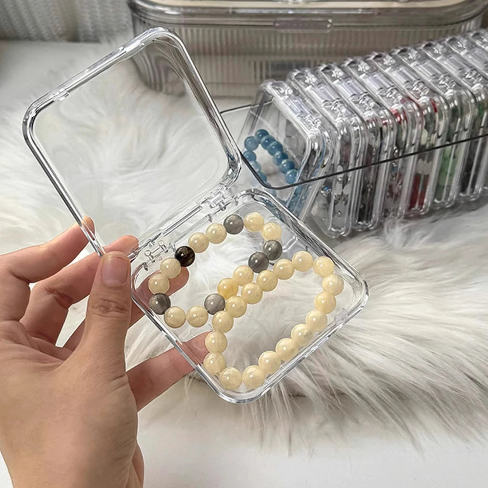 A hand holds a clear jewelry organizer box filled with light yellow and gray beads, next to a tray of clear compartments.