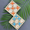 A wooden tic tac toe game set with orange and blue markers is displayed on a dark surface surrounded by green palm leaves.