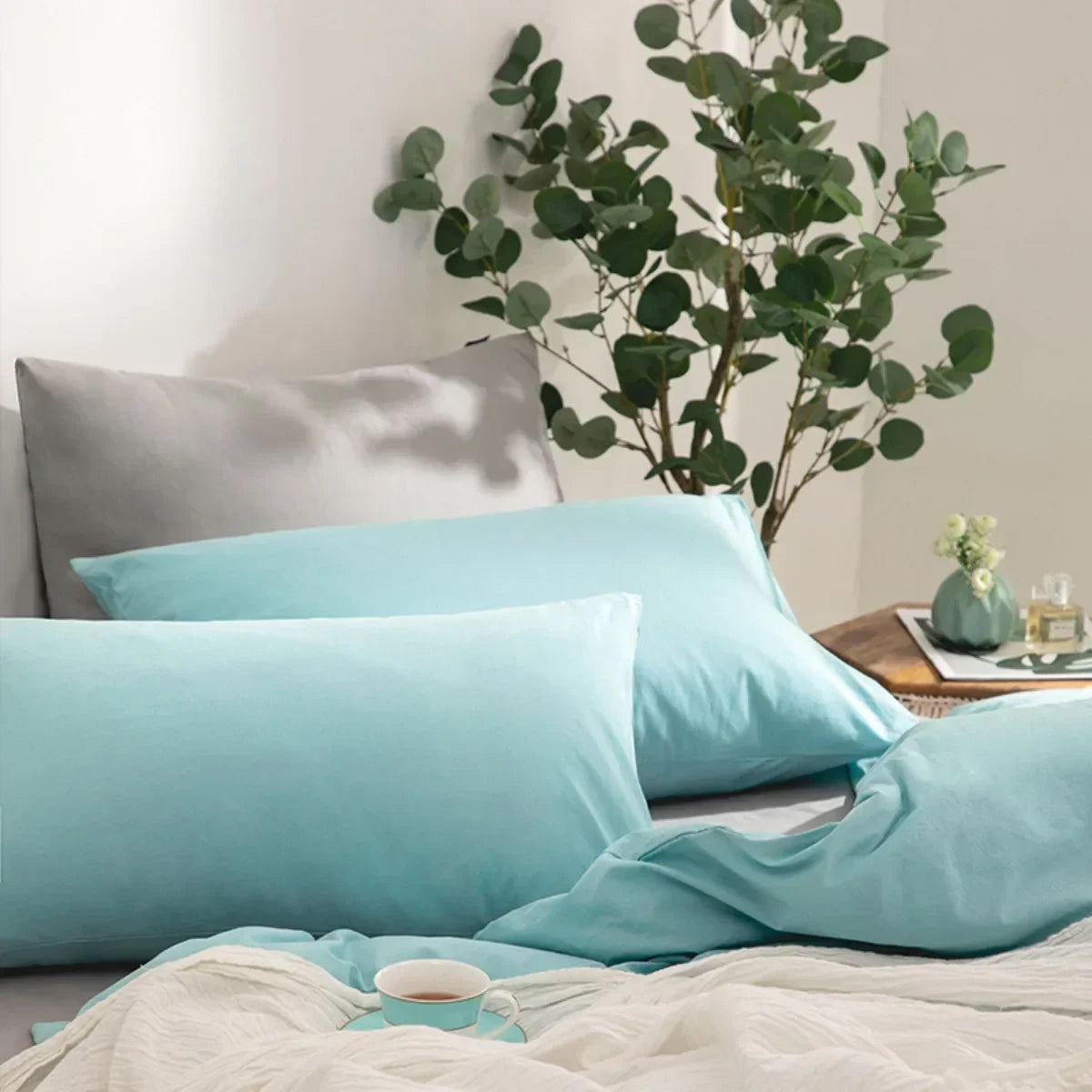 A set of four light blue microfiber pillowcases is displayed on a bed with green foliage in the background.
