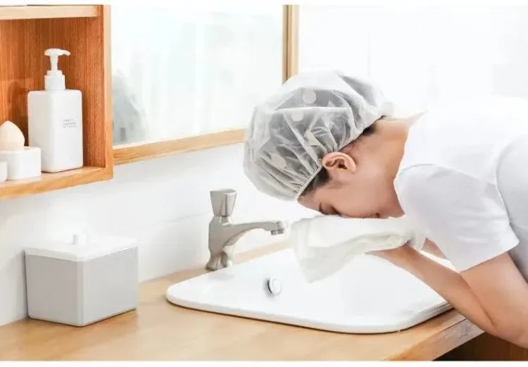 A person is using a white waterproof shower cap over their head while washing their face at a sink.