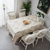 A dining table set with a cream tablecloth featuring a rustic fern print, surrounded by white wooden chairs.