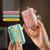 A hand holds a pink portable automatic floss pick dispenser with a swirling design and a light green version.