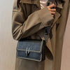 A close-up of a dark blue leather mini cross-body bag with a silver chain strap being held by a person wearing a beige trench