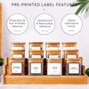 A set of five square glass spice jars with bamboo lids and labels are displayed in a wooden tray.