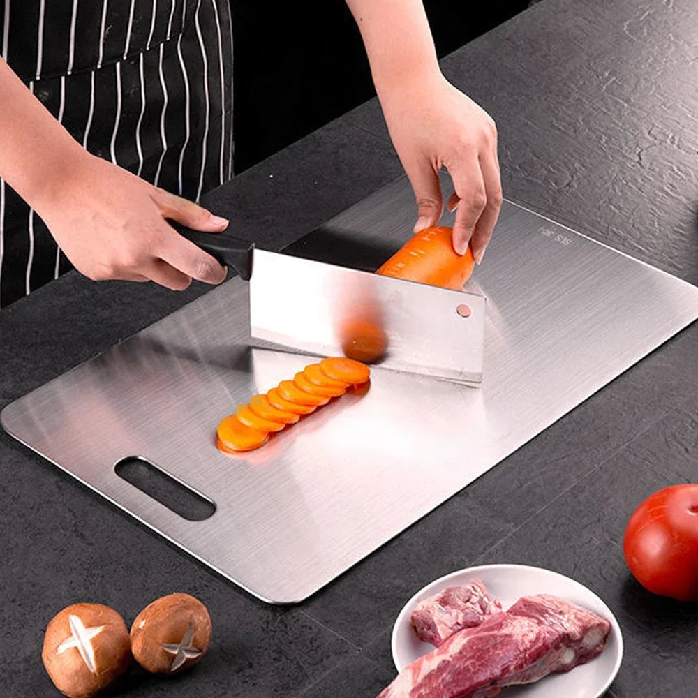 Edelstahl-Cuttingboard mit geschnittenen Karotten und rohem Fleisch auf dunkler Oberfläche.