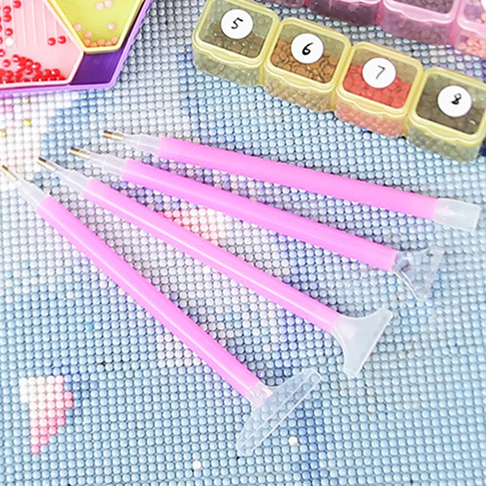A set of pink 5D diamond painting tools and a box of colored gems are displayed on a blue textured cloth.