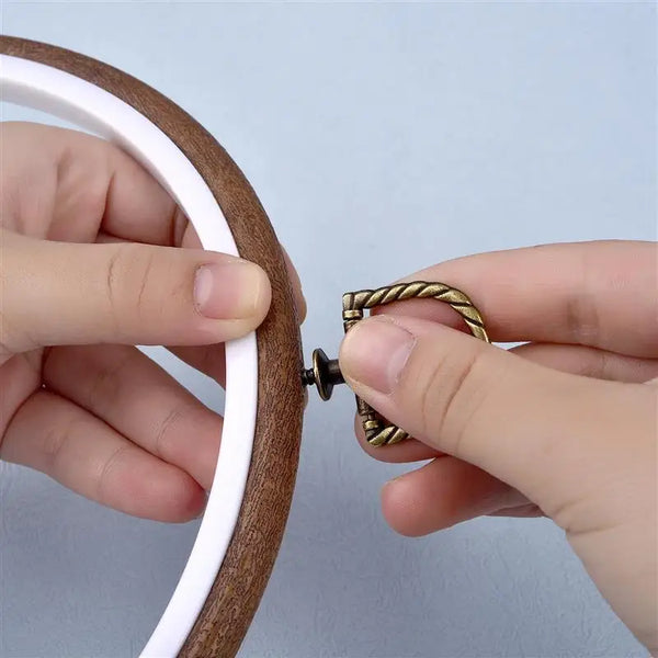 A hand is holding a wooden embroidery hoop with a white inner ring and a gold-colored metal tool.