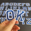 A hand holds an embroidered letter O and K from an alphabet patch set against a dark background.