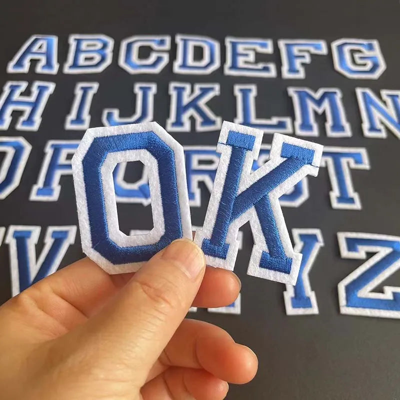 A hand holds an embroidered letter O and K from an alphabet patch set against a dark background.