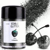 A glass jar of edible glitter powder labeled Black, next to two shiny black cherries covered in glitter.