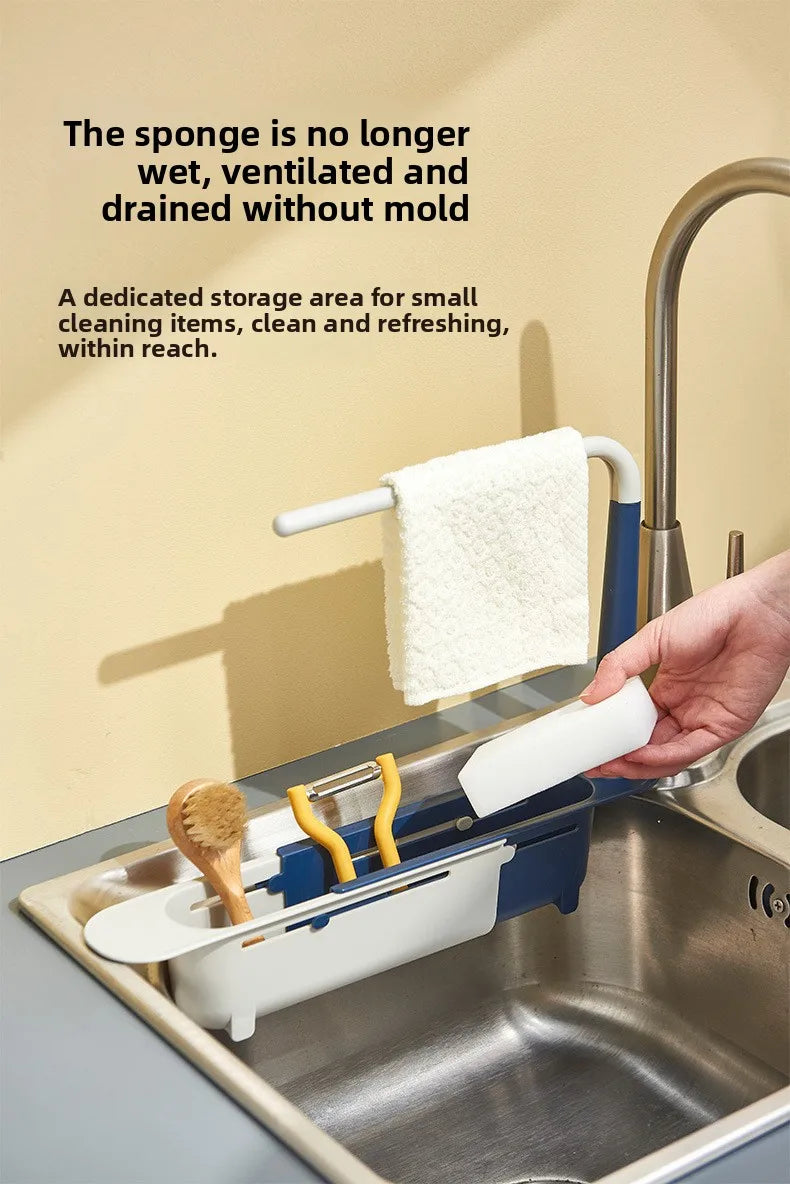 A hand places a white sponge on an adjustable sink holder with a drainer basket in a stainless steel kitchen sink.