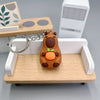 An adorable brown capybara keychain charm resting on a light wooden tray with a white rectangular box in the background.