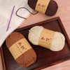 A brown crochet yarn skein and a cream-colored yarn skein are displayed in a wooden tray with a notebook and a purple pen.