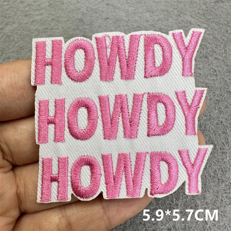 Four pink embroidered patches spelling HOWDY on a white fabric square being held in a hand.