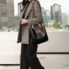 A woman wearing a tweed blazer and black pants carrying a black leather crossbody tote bag with a brown leather strap.