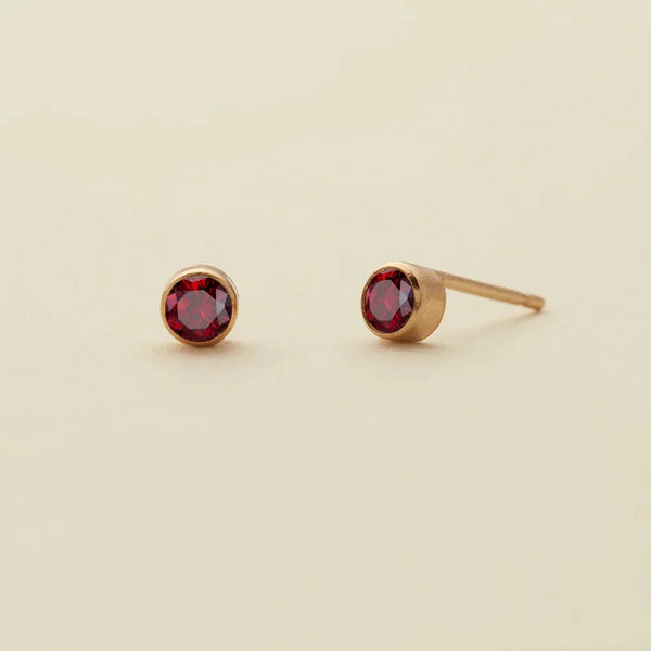 Gold-toned minimalist birthstone stud earrings featuring a deep red gemstone.