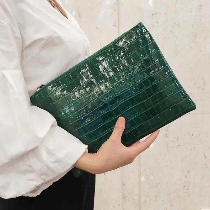 A hand holds a dark green crocodile-embossed PU leather envelope clutch against a light background.