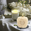 A white knitted blanket and a glowing pillar candle with a rose-shaped candle is displayed on a white cloth.