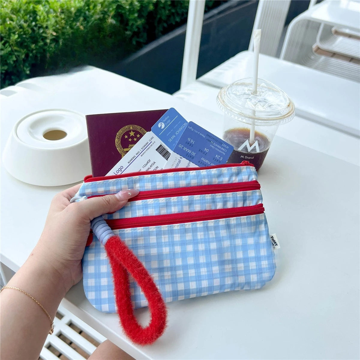 A hand holds a blue and white plaid passport holder with red trim on a white table next to a coffee cup.