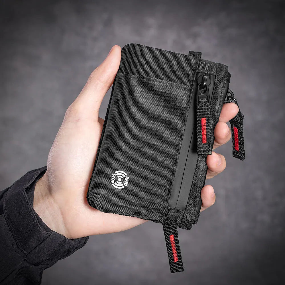 A hand holds a black RFID-blocking travel wallet with a red carabiner clip against a dark gray background.
