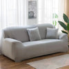 A light gray elastic stretch sofa cover for a modern couch placed in a bright room.