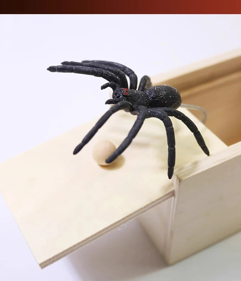 A black spider is positioned on a light wooden box, suggesting a surprise or prank item.
