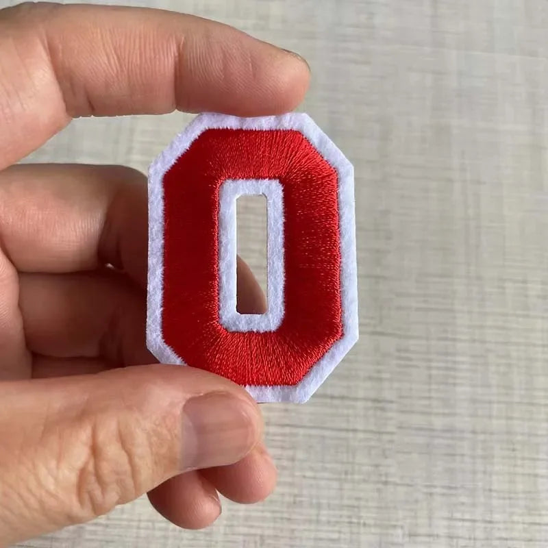 A hand holds a red iron-on 3D number patch shaped like the digit zero on a light background.