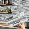 A hand is placing rectangular peel and stick waterproof tile stickers onto a gray and white subway tile backsplash.