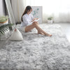 A woman sits on a fluffy gray rug reading a book next to a window with greenery.