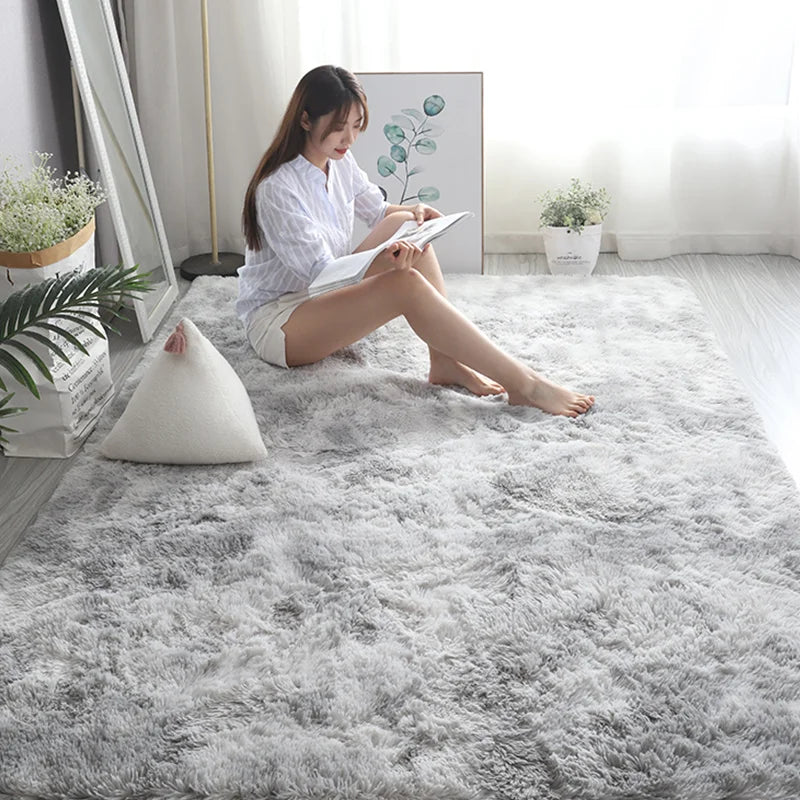 A woman sits on a fluffy gray rug reading a book next to a window with greenery.
