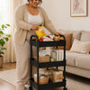 A woman is using a black 3-tier rolling utility cart with wheels to store various food items on a light rug.