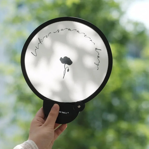 Una mano sostiene un ventilador portátil plegable negro con lente circular blanca y un diseño floral simple impreso en i