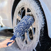 A hand is using a blue and white plush wheel cleaning brush to clean a dirty car wheel.