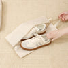 A pair of beige and tan canvas sneakers is shown inside a light brown fabric packing cube on a cream-colored textured surface