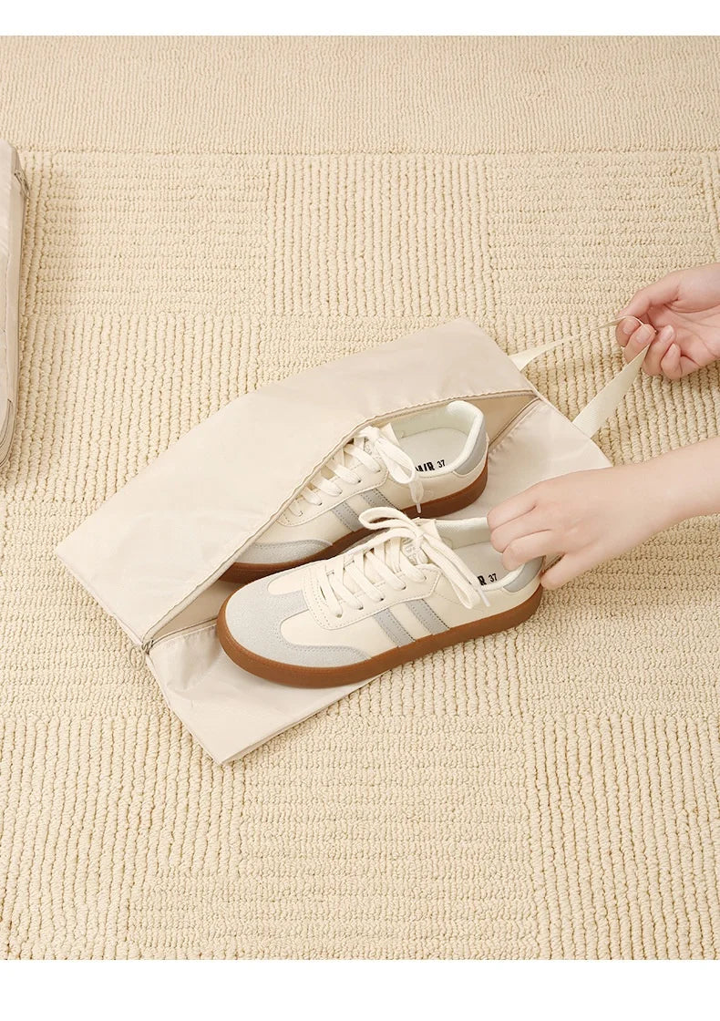 A pair of beige and tan canvas sneakers is shown inside a light brown fabric packing cube on a cream-colored textured surface