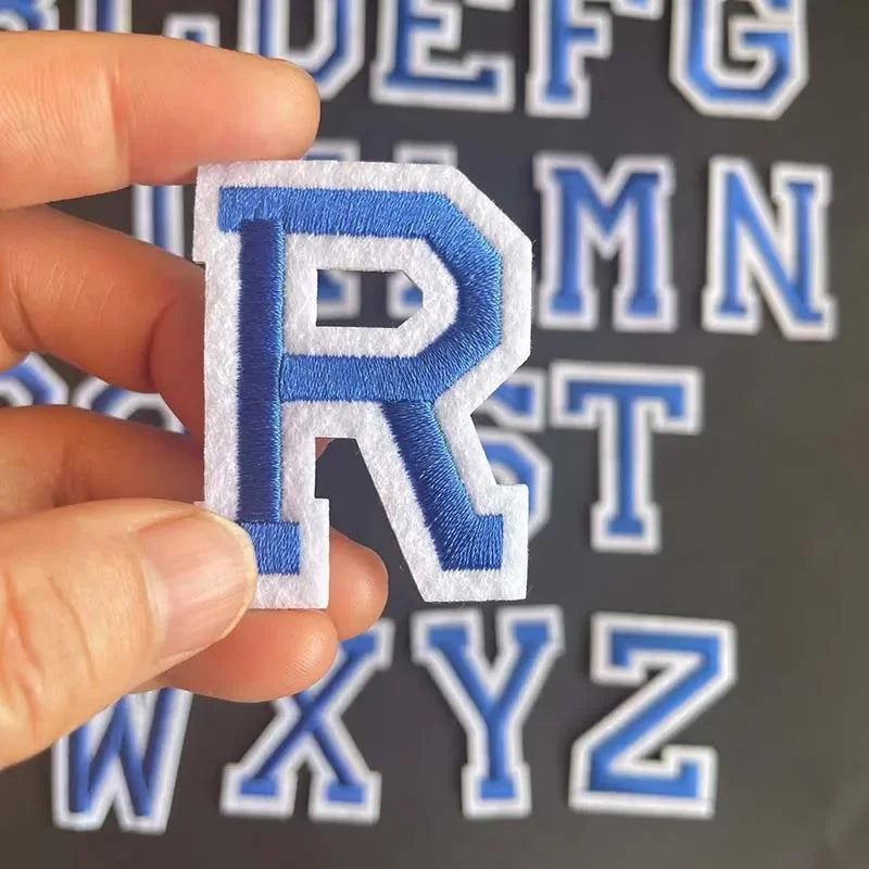 A hand holds a blue embroidered letter R from an alphabet patch set against a background of other letters.
