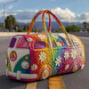 A colorful quilted travel duffle bag shaped like a vintage van with flowers and peace signs resting on asphalt.