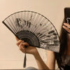 A hand holds an open black and white bamboo and silk folding fan with a floral design.